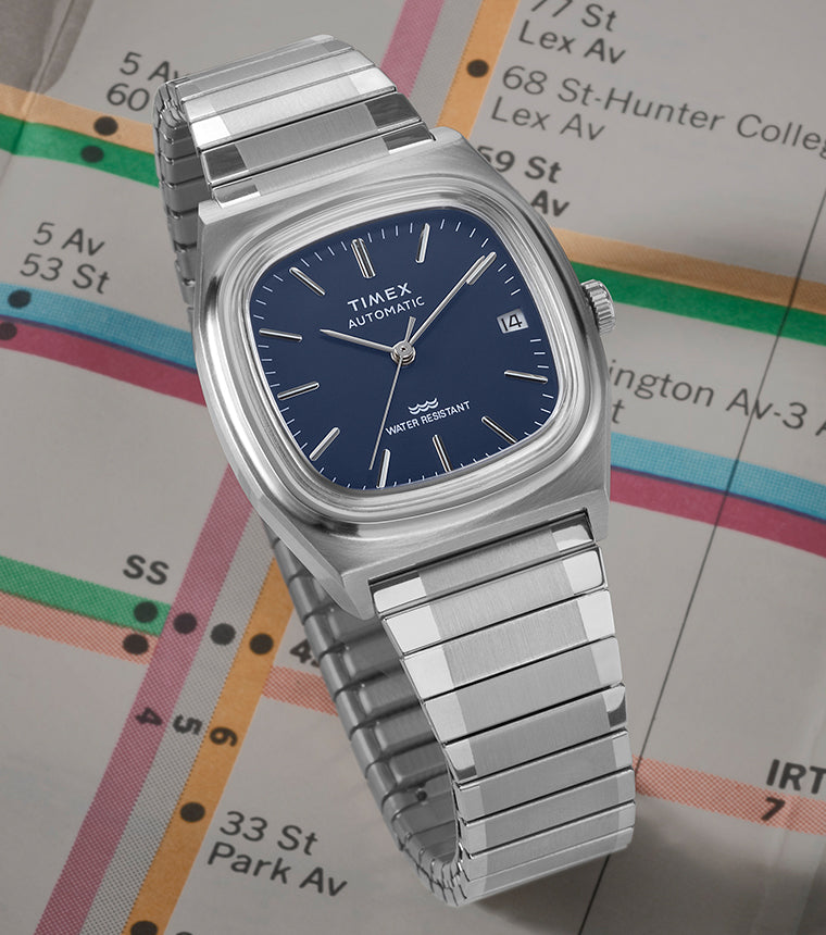 A stainless steel Timex E-Line Reissue watch with a blue dial is displayed against a vintage New York City subway map. The watch features a sleek square case, automatic movement, date display, and a water-resistant design. The subway map in the background highlights iconic NYC locations.