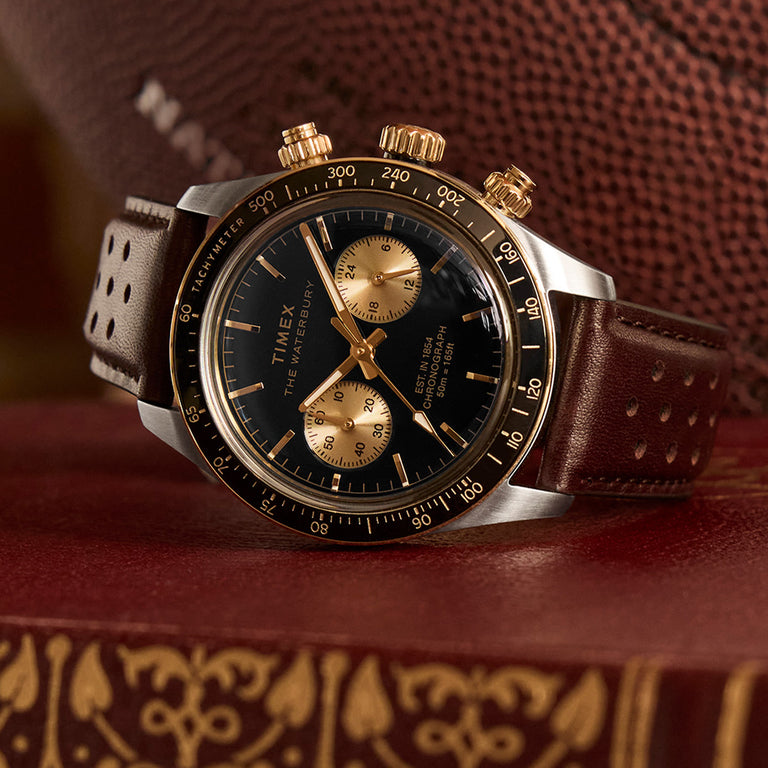 Close-up of the Timex Waterbury Chronograph watch resting on a vintage leather-bound book. The watch features a black dial with two gold sub-dials, gold accents, and a tachymeter bezel. It has a brown perforated leather strap and gold-tone pushers and crown. A textured football in the background adds a sporty, heritage-inspired touch.