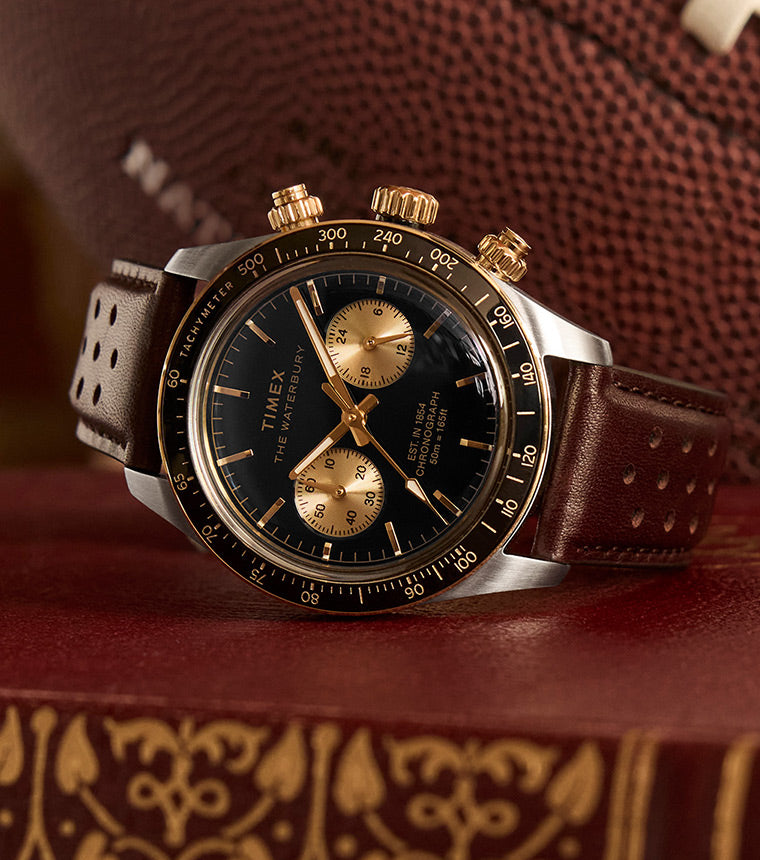 A close-up shot of the Timex Waterbury Heritage Chronograph resting on a vintage leather-bound book, with an American football in the background. The black dial with gold-tone accents, perforated brown leather strap, and polished case create a classic sports-inspired aesthetic.