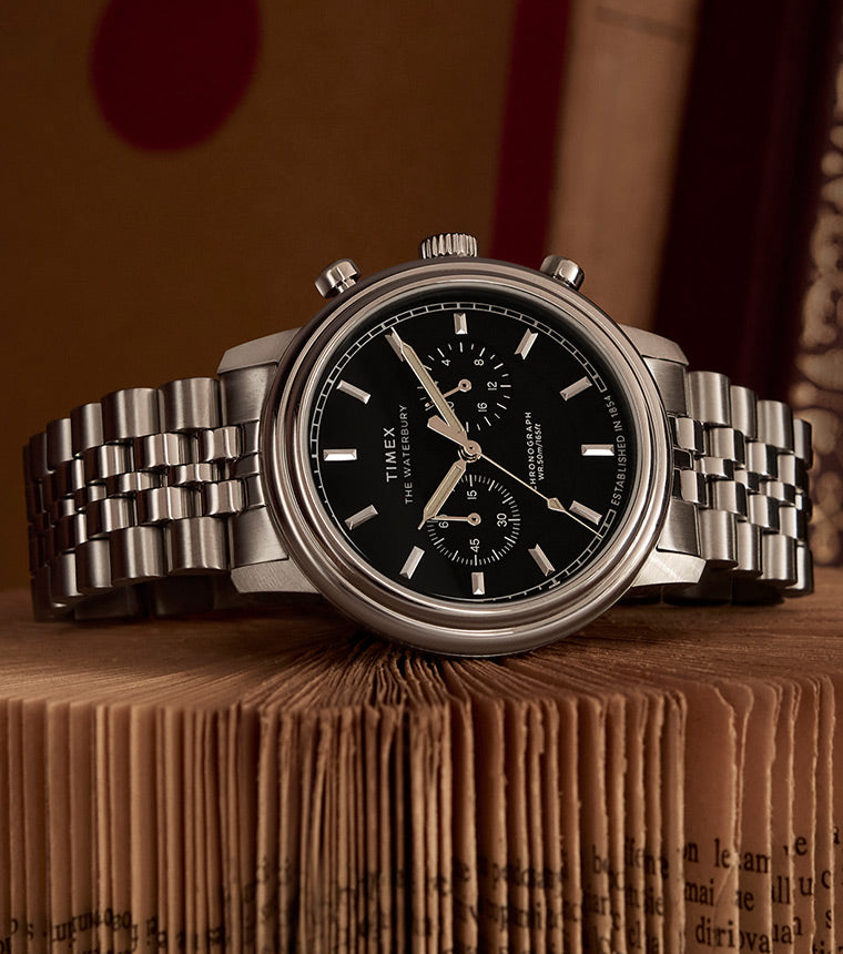 A Timex Waterbury Metropolitan Chronograph watch with a black dial, silver-tone markers, and a stainless steel bracelet, resting on a stack of aged books with intricate bindings in the background.