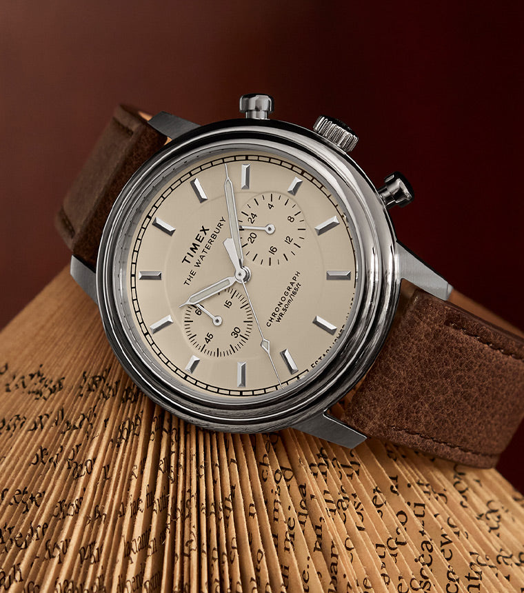 A Timex Waterbury Metropolitan Chronograph watch with a cream-colored dial, silver-tone markers, and a brown leather strap, elegantly placed on the fanned pages of an aged book against a warm-toned background.