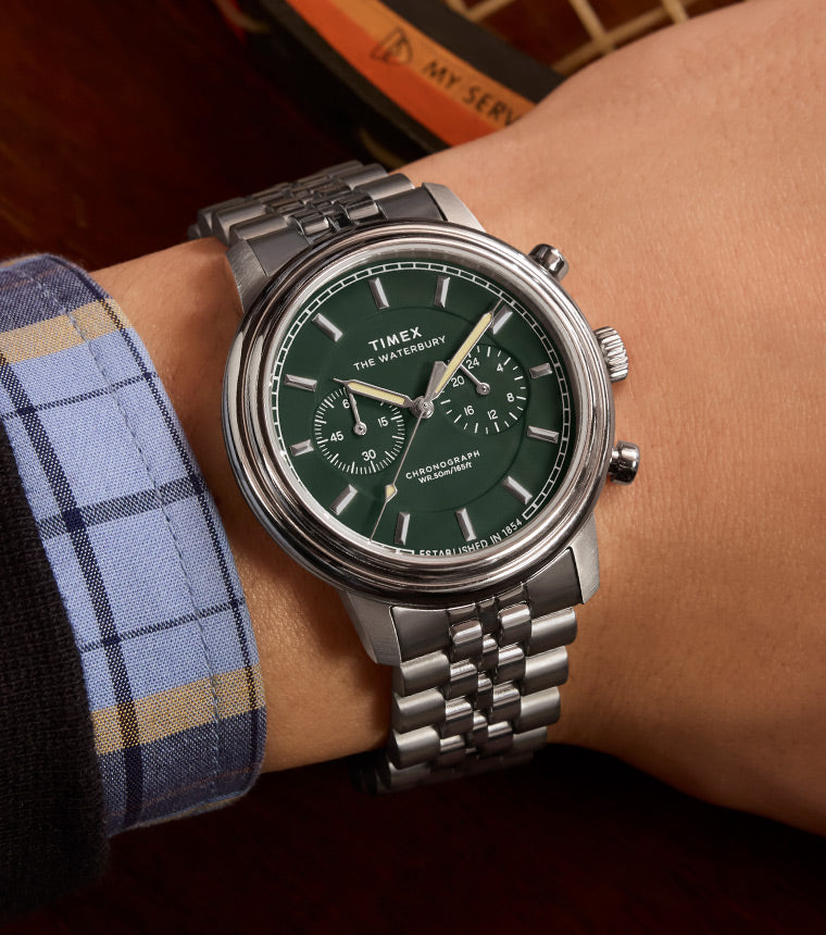 A Timex Waterbury Metropolitan Chronograph watch with a green dial, silver-tone markers, and a stainless steel bracelet, worn on a person’s wrist with a vintage wooden tennis racket in the background.
