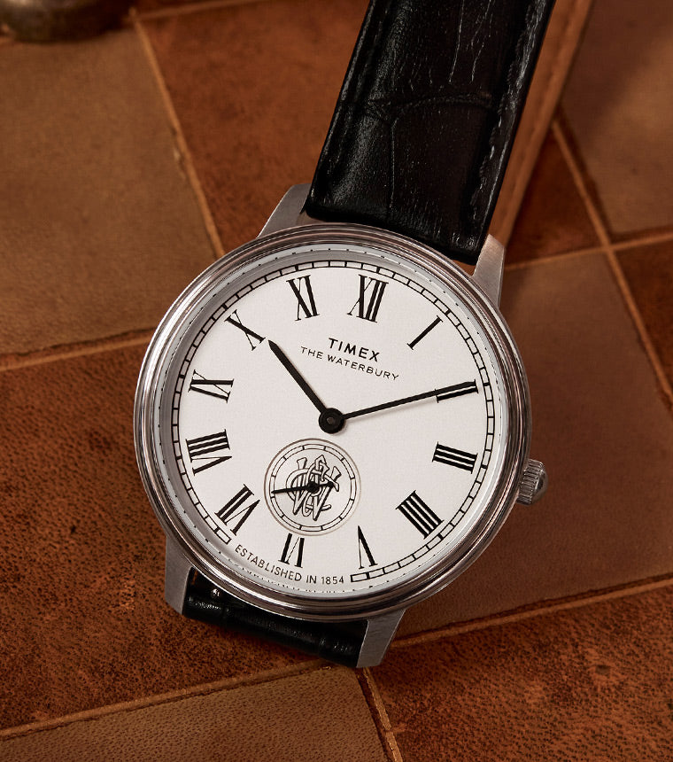 A close-up of a Timex Waterbury Metropolitan Sub-Second wristwatch featuring a white dial, black Roman numeral hour markers, and a black leather strap laid perfectly against brown tiles.