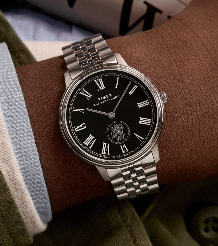 A close-up of a Timex Waterbury Metropolitan Sub-Second wristwatch with a black dial, silver Roman numeral hour markers, and a stainless steel bracelet, worn by a person dressed in a green jacket and white cuff. A newspaper is partially visible in the background.