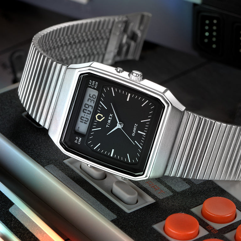 Timex Q Analog-Digital watch with a sleek rectangular case and black dial, placed atop a retro gaming console controller. The watch features dual display—an analog dial with minimalist hour markers and a digital screen displaying the time. It is paired with a ribbed stainless steel bracelet, evoking a vintage tech aesthetic.