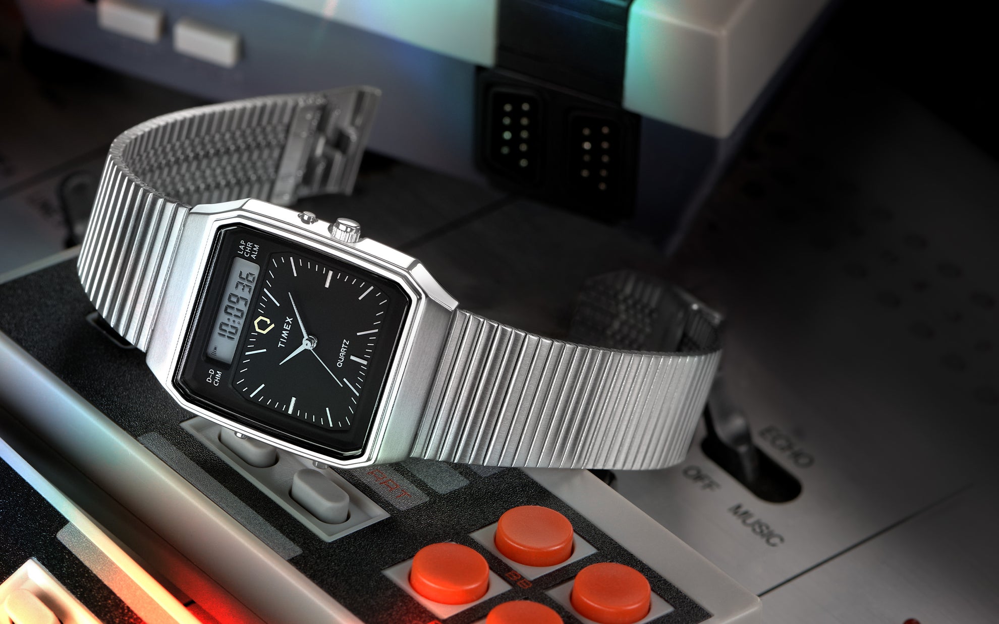 A Q Timex® 1982 Ana-Digi Reissue watch with a black dial and silver-tone case is placed on a retro gaming console. The watch has an analog and digital hybrid display, a stainless steel bracelet, and a distinctive vintage aesthetic. The background features an old-school controller with red buttons and a nostalgic gaming atmosphere.