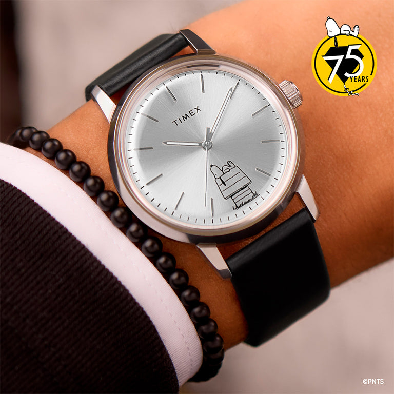 Timex wristwatch with a silver sunburst dial and black leather strap, featuring a minimalist Snoopy lying on his doghouse at the 5 o'clock position. Peanuts 75th anniversary badge shown in the upper-right corner.