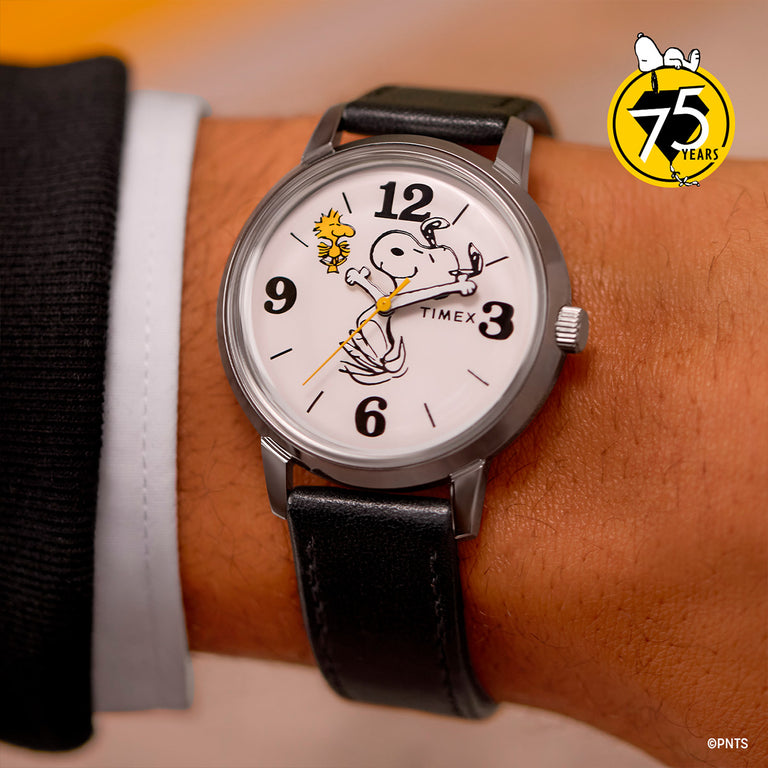 Timex analog watch with a white dial, bold black numerals, and black leather strap. Features Snoopy dancing with Woodstock, with their arms as the hour and minute hands. Peanuts 75th anniversary badge displayed in the top-right corner.