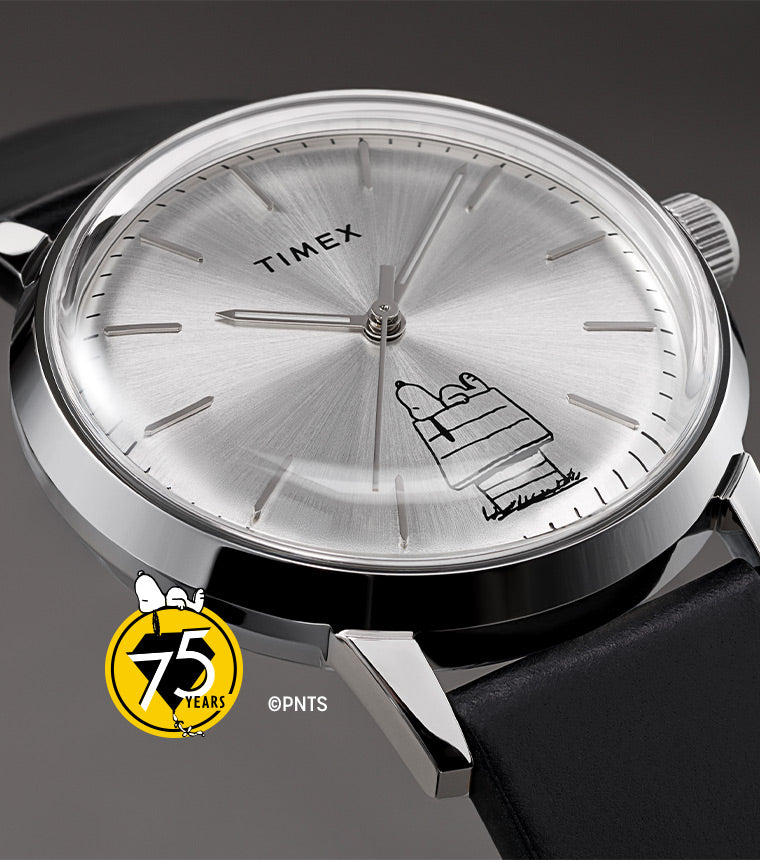 A close-up of the Timex x Peanuts® Marlin® Automatic watch with a silver dial, black leather strap, and an illustration of Snoopy lying on his doghouse. The stainless steel case and minimalist design are showcased against a neutral background. A small “75 Years” Peanuts® logo is visible.