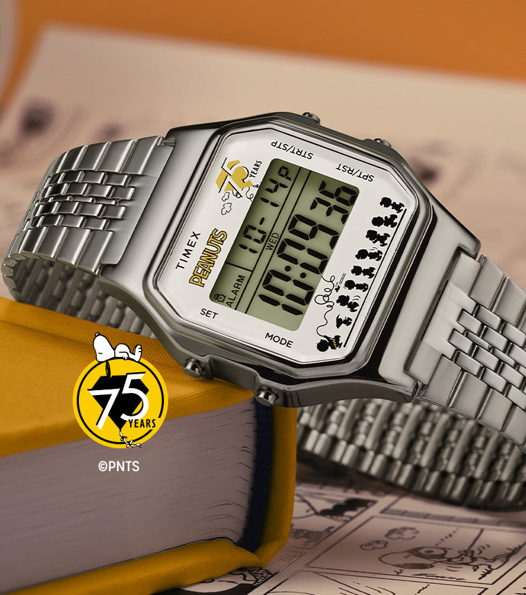 A close-up of the Timex x Peanuts® T80 digital watch, placed on a yellow hardcover book with Peanuts® comic strip pages in the background. The watch features a silver-tone stainless steel bracelet, a digital display with a “75 Years” Peanuts® logo, and a row of silhouetted Peanuts® characters walking along the bottom.