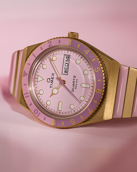 Timex Q watch with a pink dial, gold-tone case, and pink bezel, featuring a day-date display and a two-tone pink and gold bracelet.