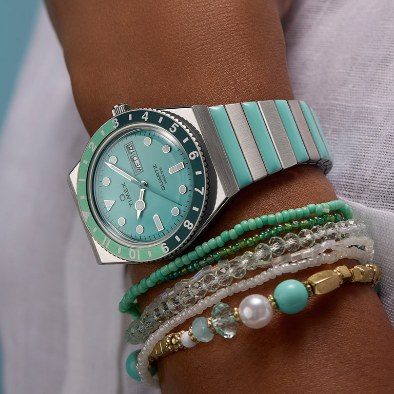 Close-up of the Q Timex® watch on a wrist, featuring a pastel aqua dial, matching day-date window, and a two-tone bezel in black and mint green. The stainless steel bracelet has alternating aqua accents, complementing the stacked beaded bracelets in green, white, and gold tones. The soft, stylish look reflects a fresh, retro-inspired aesthetic with a modern twist.