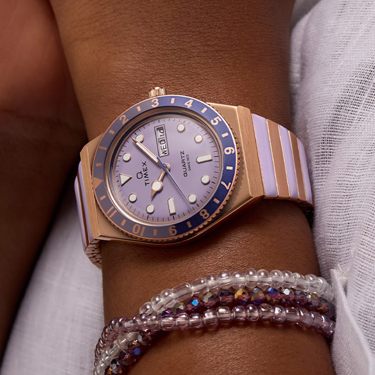 A close-up of a wrist wearing a Timex Q women’s quartz watch with a rose gold-tone case, lavender dial, and two-tone lavender and rose gold expansion band. The watch features a rotating navy and gold bezel, round indices, and a day-date window at 3 o’clock. The wearer also has layered beaded bracelets in shades of purple and clear, against a white textured fabric background.