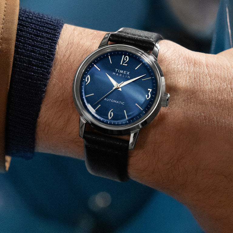 Timex Marlin Automatic watch with a sunburst blue dial, silver-tone case, and black leather strap, shown on a wrist. The dial features silver hour markers with numerals at 12, 3, 6, and 9, combining a vintage-inspired design with modern elegance.