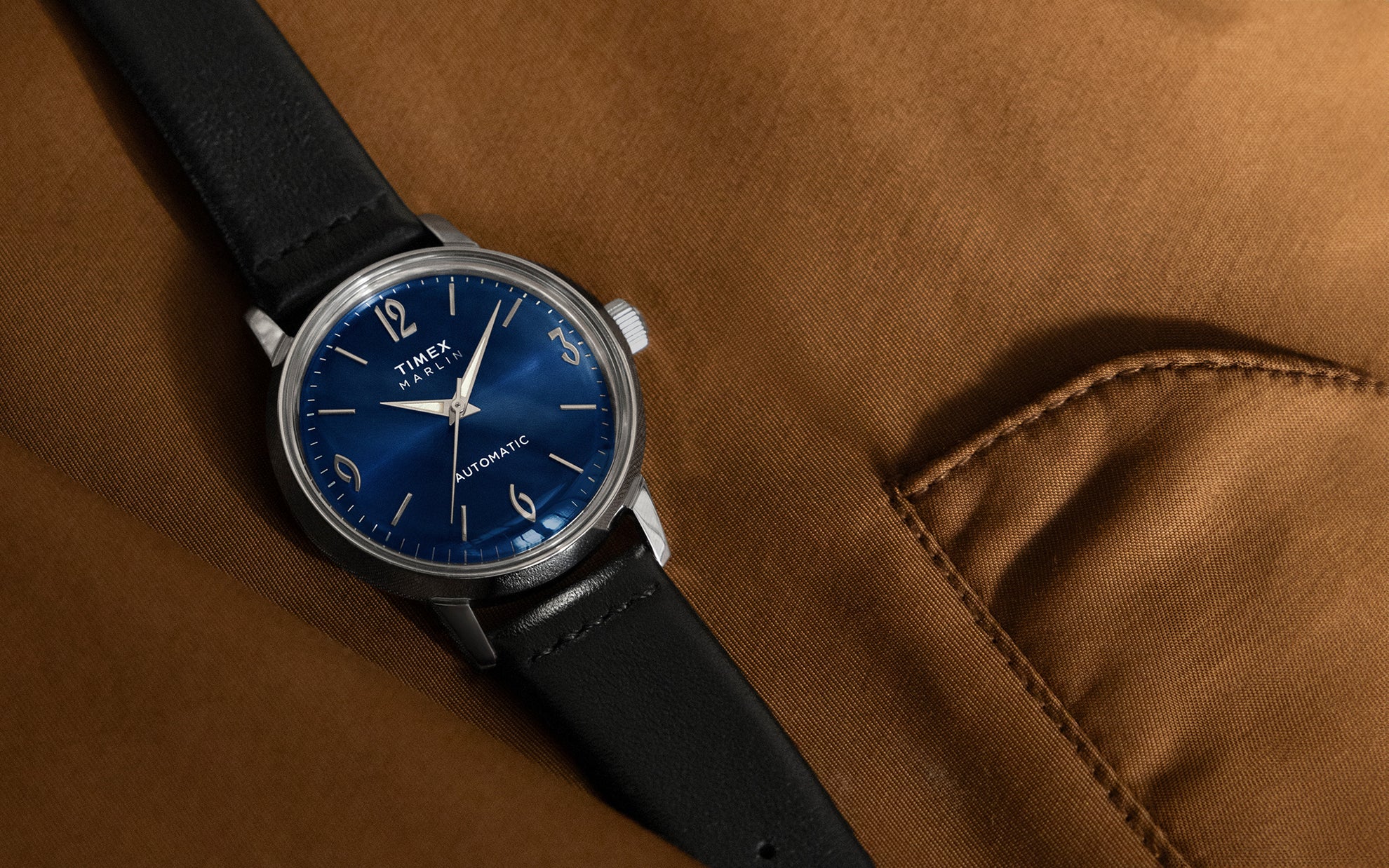 A sophisticated automatic watch with a deep blue dial, silver indices, and numerals at 12, 3, 6, and 9 o’clock. The case is stainless steel, paired with a black leather strap, elegantly placed on a brown fabric background.