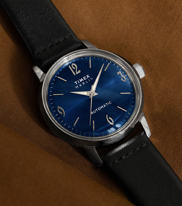 A sophisticated automatic watch with a deep blue dial, silver indices, and numerals at 12, 3, 6, and 9 o’clock. The case is stainless steel, paired with a black leather strap, elegantly placed on a brown fabric background.