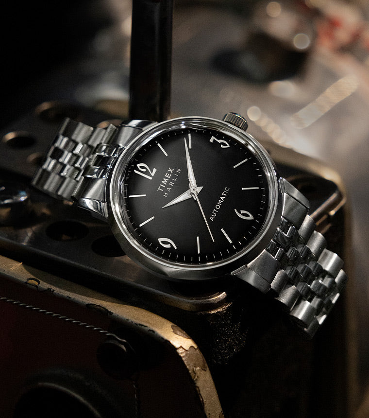 A refined automatic watch featuring a black sunburst dial, silver-tone numerals at 12, 3, 6, and 9 o’clock, and a stainless steel case. The watch is paired with a polished stainless steel bracelet and is placed on an industrial metal surface.