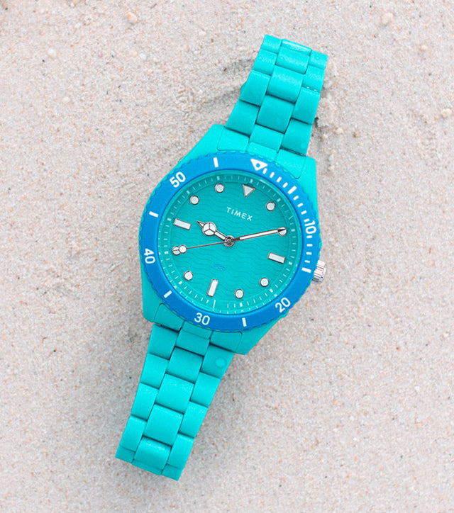 A turquoise Timex Legacy #tide™ watch placed on soft sand near a coral rock, showcasing its ocean-inspired design and sustainable materials made from upcycled ocean-bound plastic.
