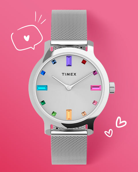 A stylish Timex watch with a silver mesh band and a clean white dial, accented by colorful gemstone hour markers in rainbow shades. The background is soft pink with hand-drawn hearts and a speech bubble.