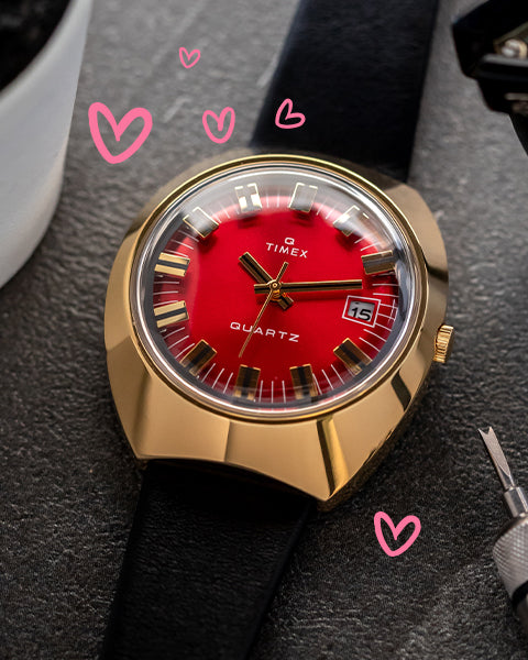 A close-up of a Timex quartz watch featuring a bold red dial, gold-toned case, and black leather strap. The watch face includes a date display and gold hour markers. The background has a textured surface, with doodled pink hearts adding a romantic touch.