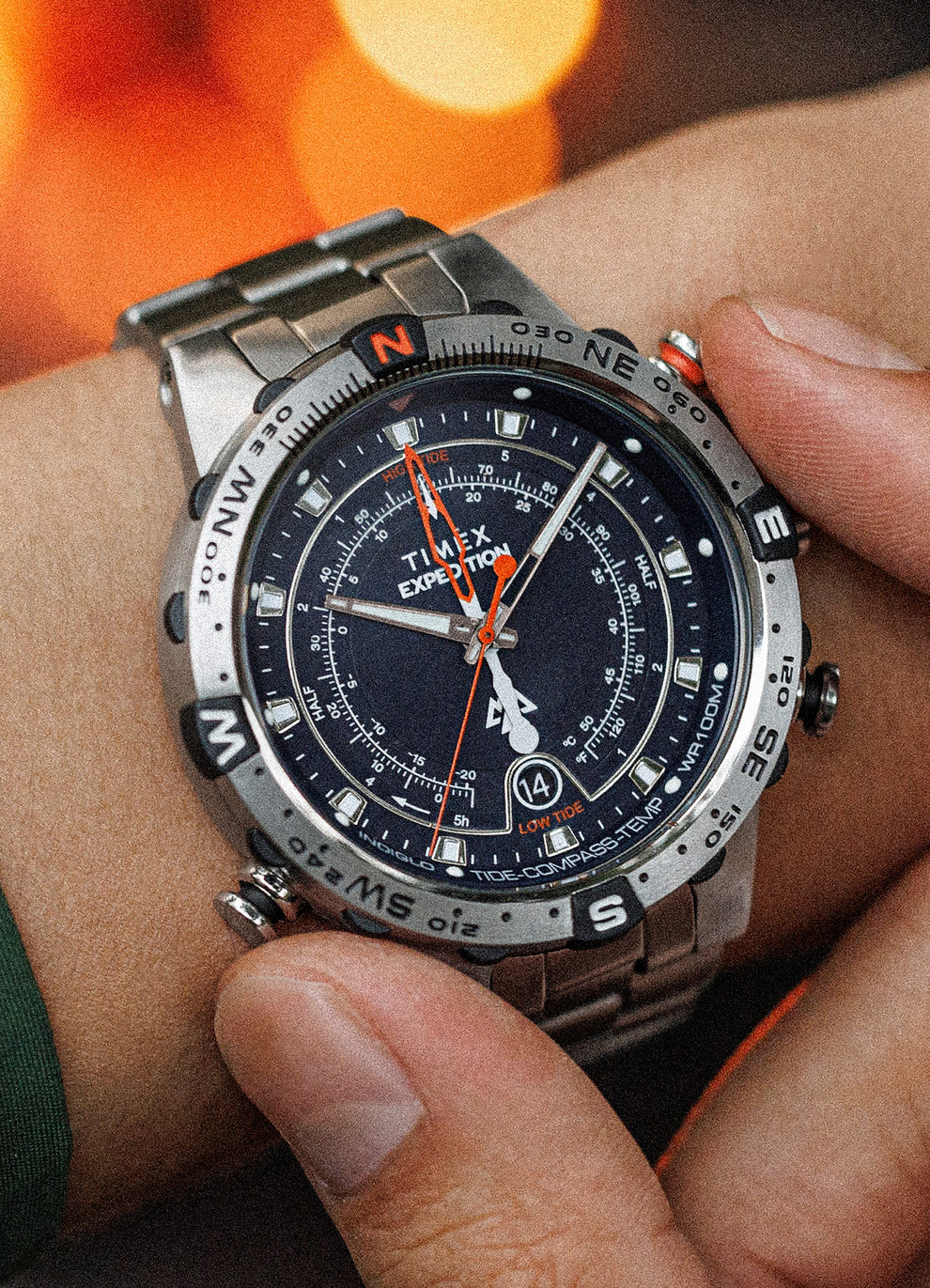 Close-up of a person adjusting the crown of the Timex Expedition Tide-Temp-Compass watch while wearing it. The stainless steel timepiece showcases a black dial with compass markings, tide and temperature indicators, and luminous hands.