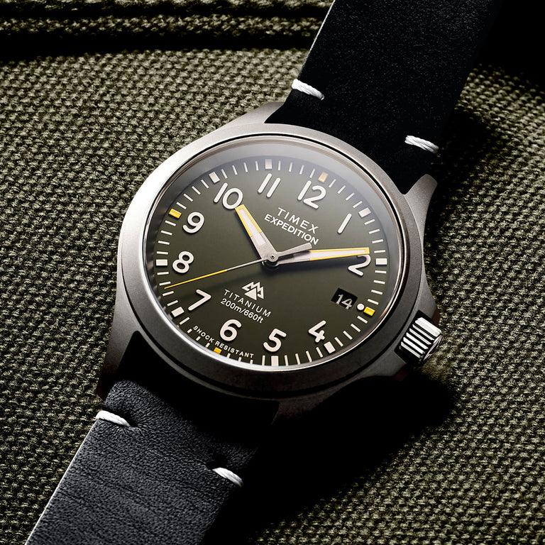 A close-up of a Timex Expedition watch resting on a textured olive-green fabric background. The watch features a matte titanium case with a dark green dial, bold white Arabic numerals, and luminous hands with yellow accents. It has a date window at the 3 o’clock position and is labeled “Titanium 200m/660ft” and “Shock Resistant.” The black leather strap with white stitching adds a rugged and adventure-ready feel to the design, reinforcing its durability and field-inspired aesthetic.