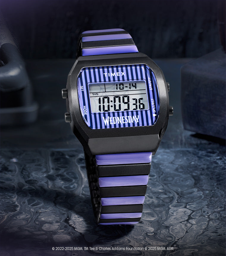 A moody flatlay image of the Timex x Wednesday T80 digital watch. The watch features a black and purple striped resin strap and a digital display framed by a bold striped dial with the word "WEDNESDAY" prominently displayed. It rests on a dark, textured surface with cobweb-covered vintage items and gothic books in the background, evoking the eerie aesthetic of the "Wednesday" series.