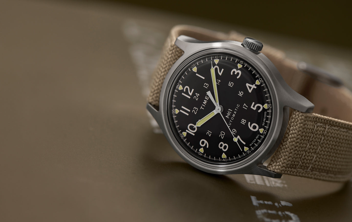 Timex® MK1® Automatic watch with a brushed gray case, black field dial, and tan fabric strap, displayed at an angle on a muted olive surface.