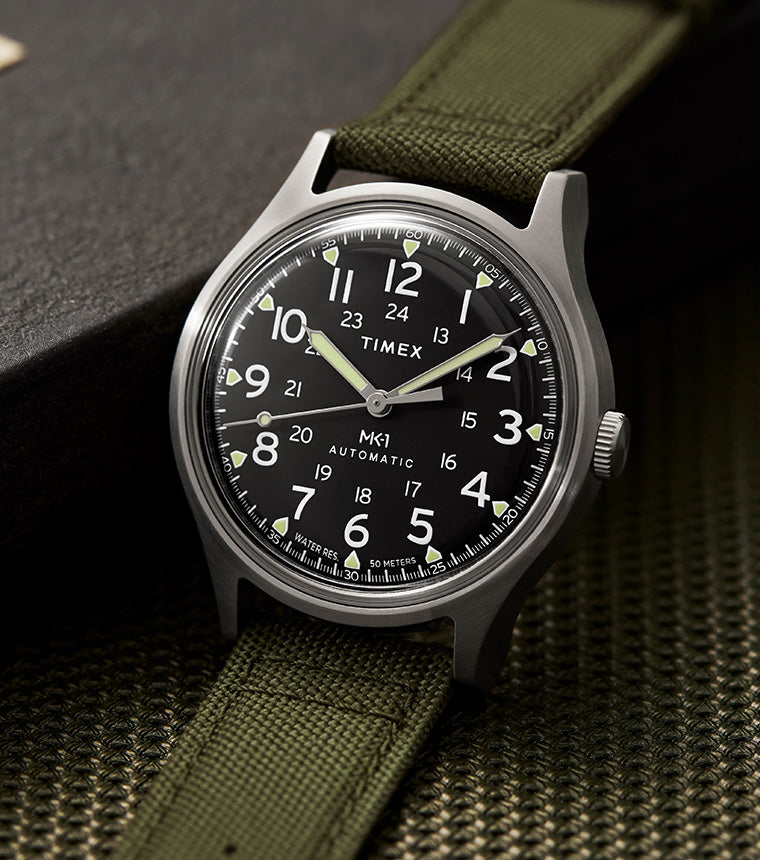 A Timex MK1® Automatic field watch with a black dial, white Arabic numerals, and luminous hands, displayed on an olive green fabric strap. The watch has a stainless steel case and is set against a textured dark background with a metal nameplate. The design reflects a military-inspired aesthetic with clear, easy-to-read markings and a rugged yet stylish appearance.