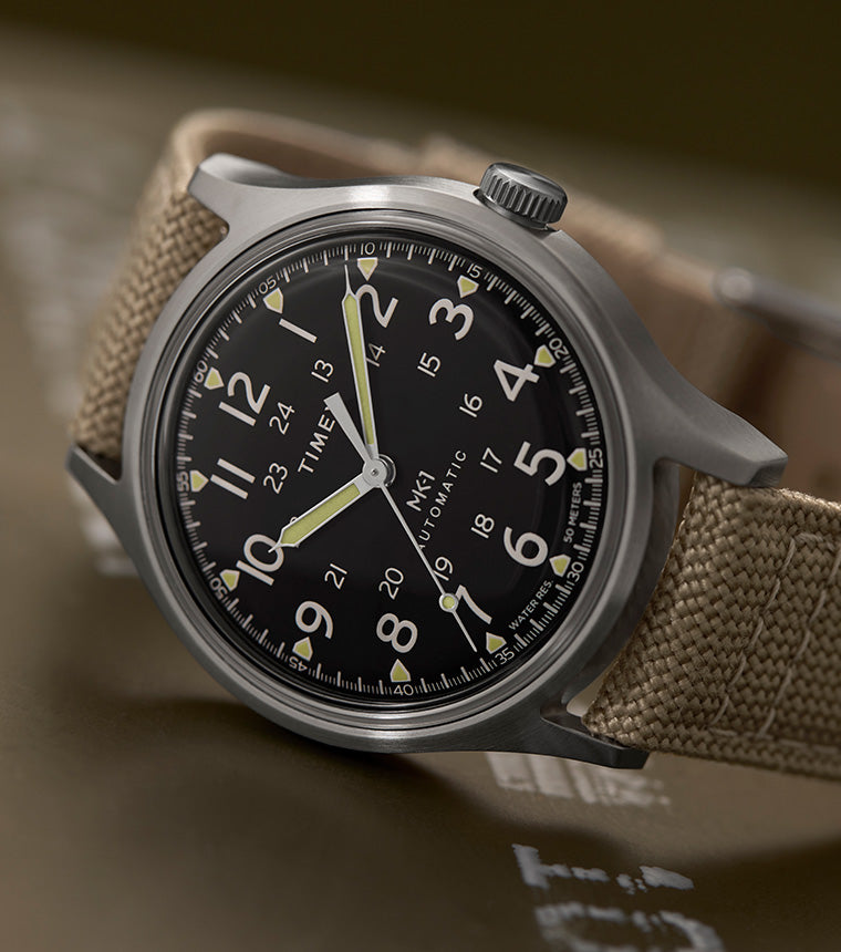 Timex® MK1® Automatic watch with a brushed gray case, black field dial, and tan fabric strap, displayed at an angle on a muted olive surface.