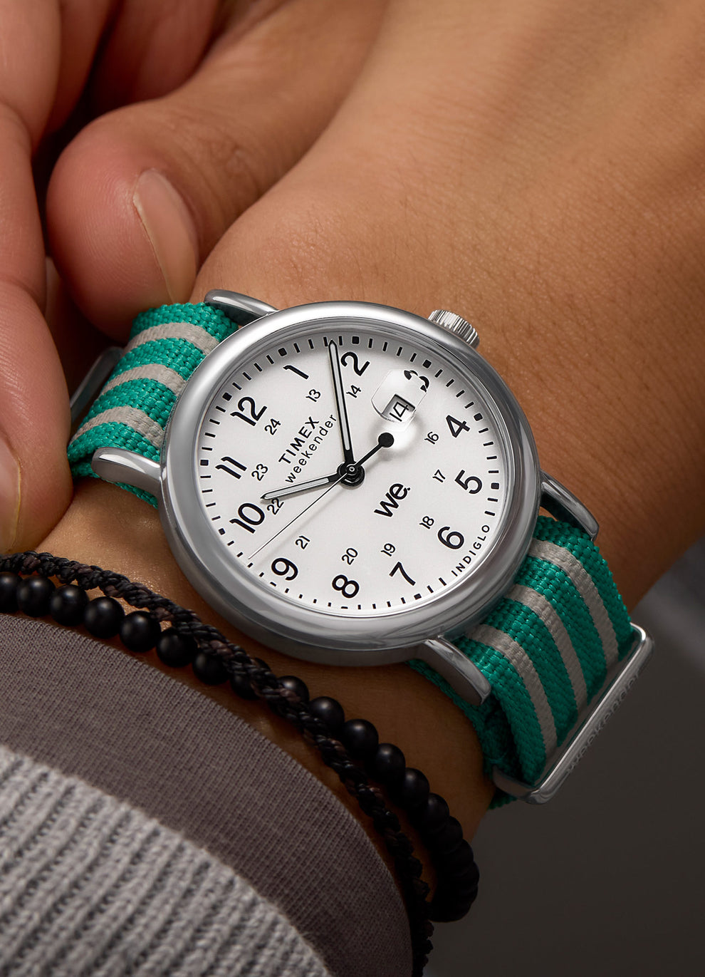 Close-up wrist shot of a Timex Weekender with an aqua-and-gray striped strap, styled with a black and brown beaded bracelet.