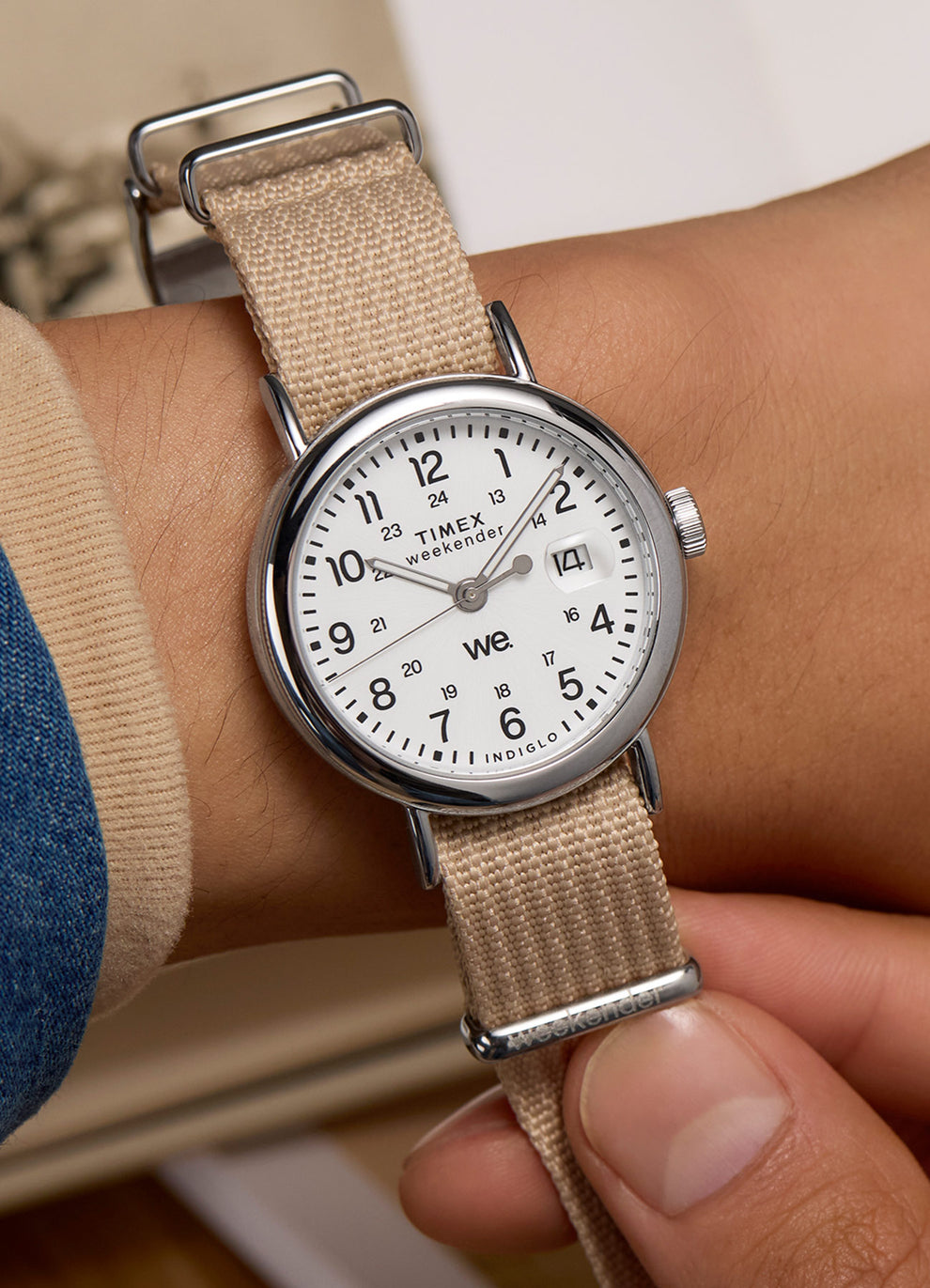 A close-up of the Timex Weekender 37mm on wrist, showcasing its casual beige woven strap, white dial with black numerals, and Indiglo functionality, paired with a denim jacket and tan sweater.