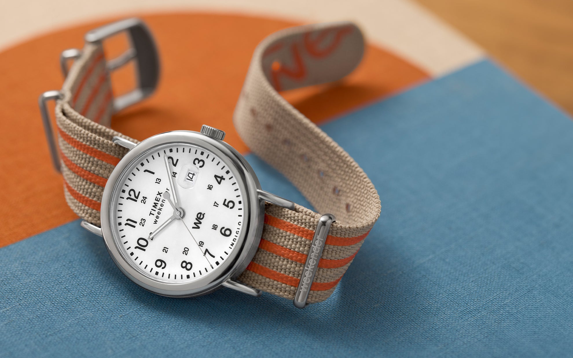 Timex Weekender laid on a textured surface with color blocks in orange and blue, featuring a beige and orange striped strap.