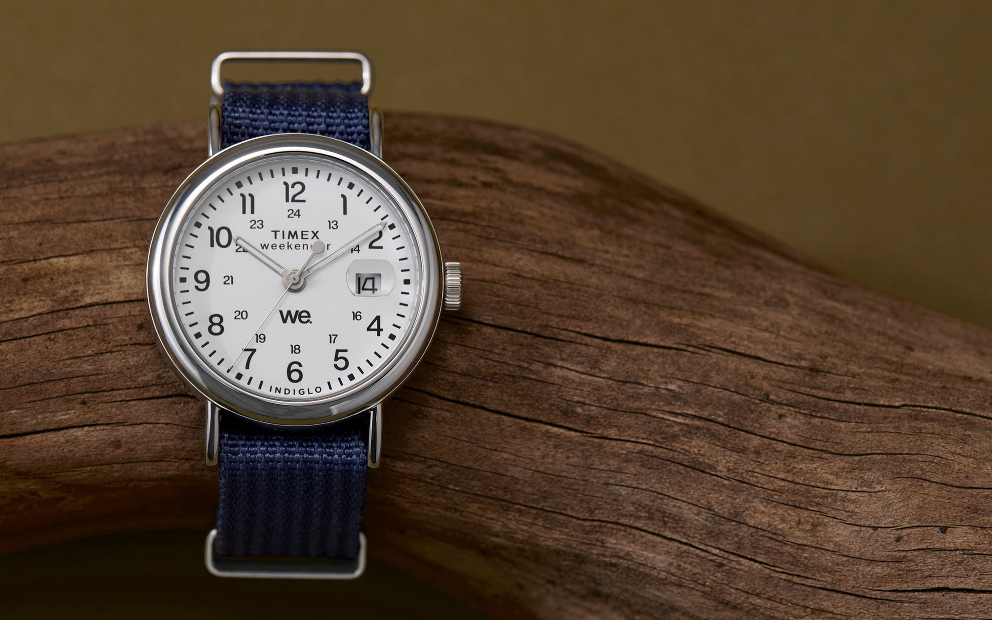 The Timex Weekender 37mm watch resting on a curved driftwood piece, featuring a white dial, black Arabic numerals, and a navy-blue fabric strap.