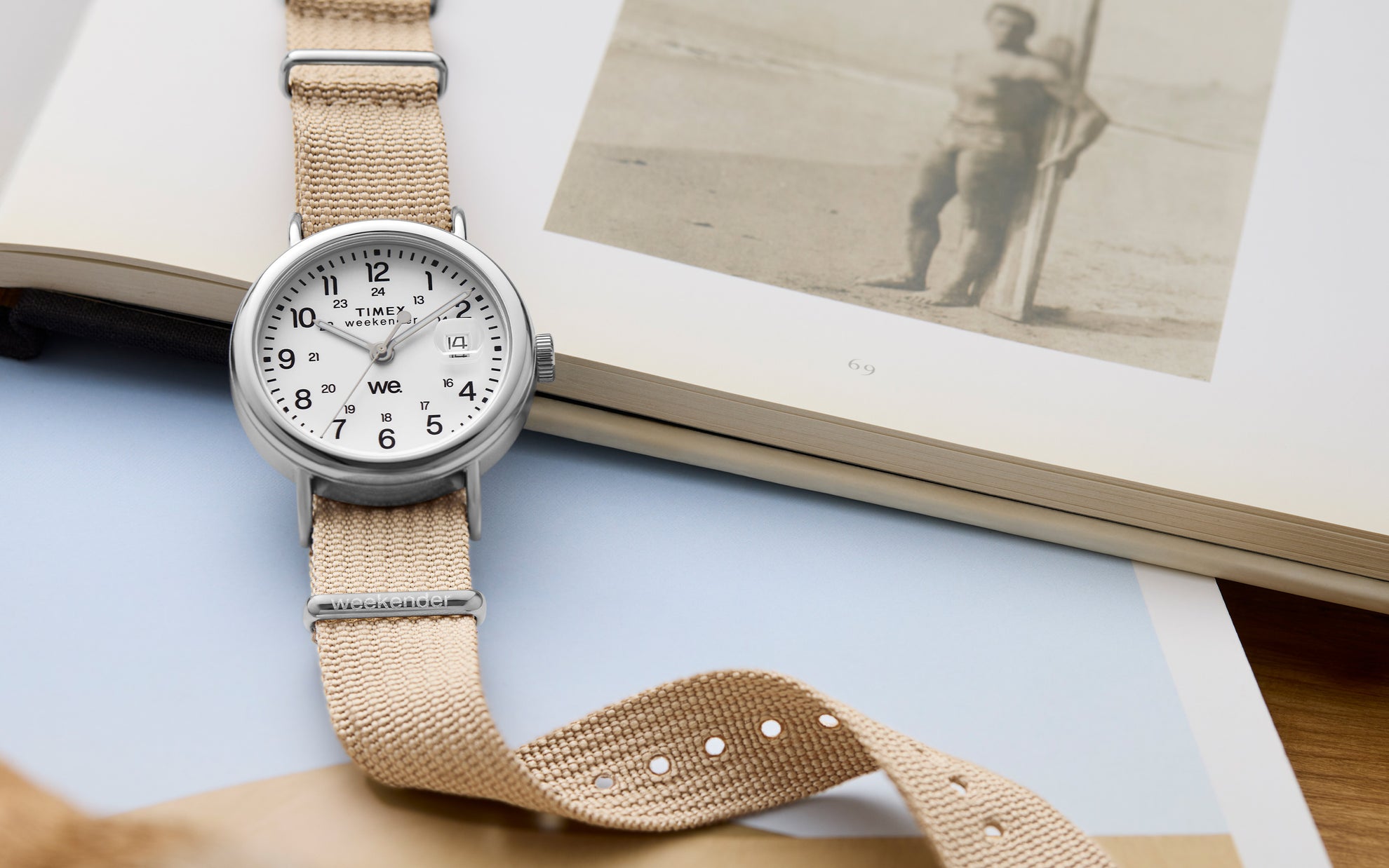 A Timex Weekender 37mm watch with a clean white dial and black numerals, displayed on an open book. It features a beige fabric strap and polished silver-tone case, resting across a vintage photograph and a pastel blue sheet.