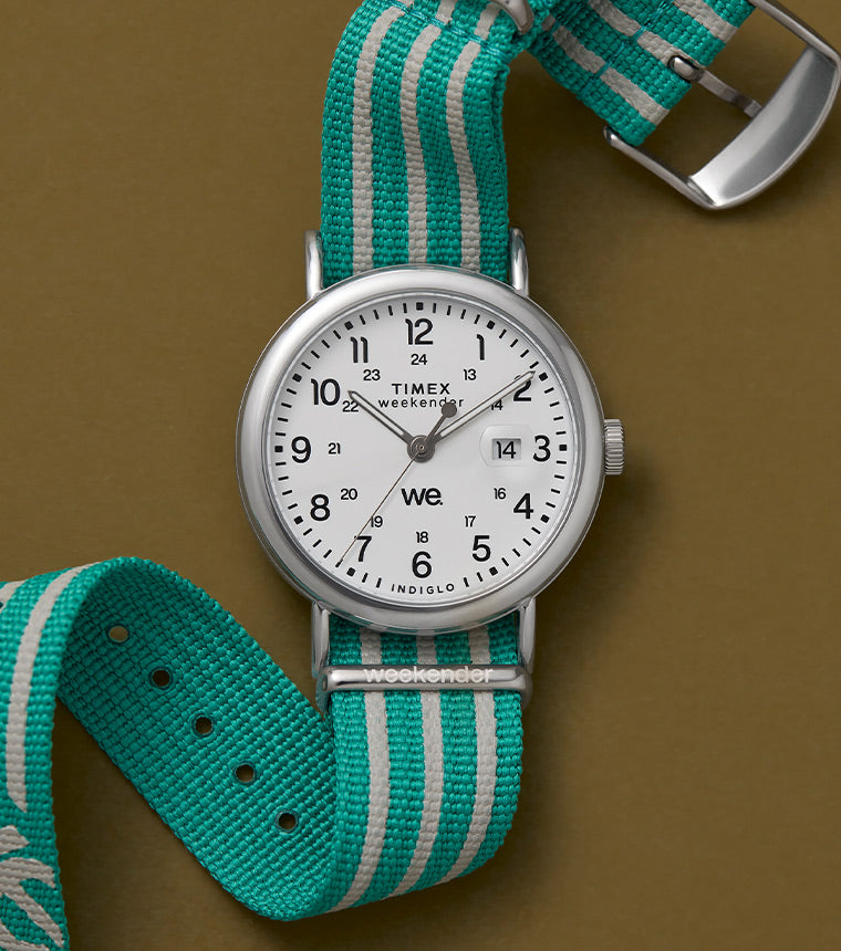 A Timex Weekender watch laid flat against a khaki background, featuring a white dial and an aqua-and-gray striped fabric strap displaying the text "weekender".