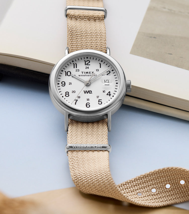 A Timex Weekender 37mm watch with a clean white dial and black numerals, displayed on an open book. It features a beige fabric strap and polished silver-tone case, resting across a vintage photograph and a pastel blue sheet.