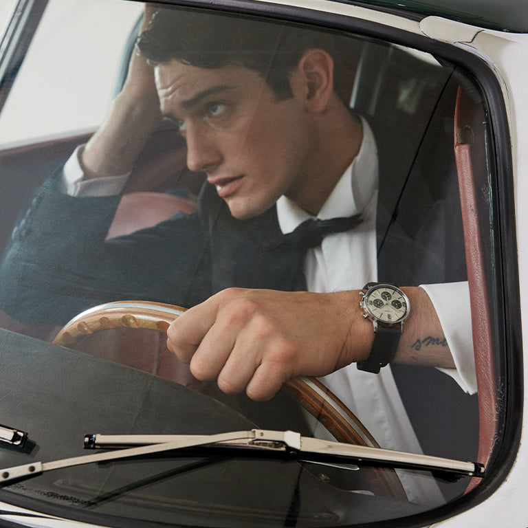 A man in a tuxedo sits inside a classic car, gripping the wooden steering wheel with one hand and leaning his head on the other in a thoughtful pose. Seen through the windshield, he wears a black leather strap chronograph watch with a cream dial and dark subdials. The dramatic lighting and formal attire give a cinematic, vintage feel.
