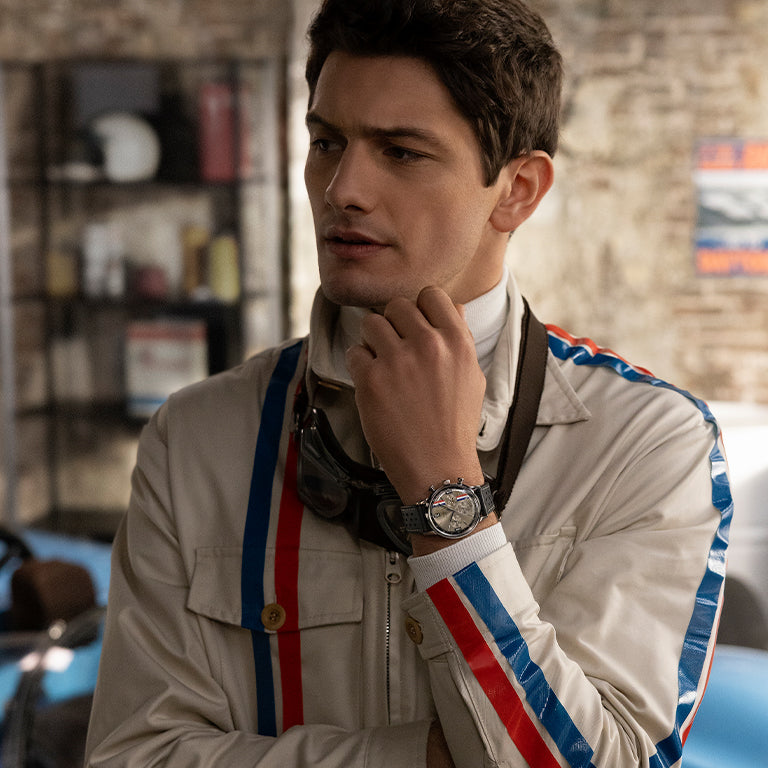 A man in a retro-inspired racing jacket with red and blue stripes poses thoughtfully in a garage or workshop setting. He wears a chronograph watch with a rugged design, evoking motorsport heritage.