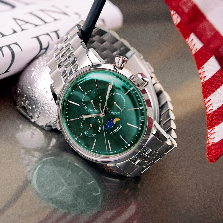 A close-up shot of a sophisticated Timex wristwatch with a deep emerald green dial, silver indices, and multiple subdials, including a moon phase complication. The watch features a polished stainless steel bracelet and is resting elegantly atop a glossy surface next to a rolled-up newspaper and a red-and-white striped fabric, evoking a sense of refined lifestyle and attention to detail.