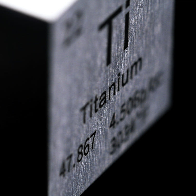 A macro shot of a metallic surface engraved with the chemical symbol "Ti" and the word "Titanium," along with its atomic number and weight. The brushed metal texture is prominently displayed, emphasizing the material's durability and sleek appearance.