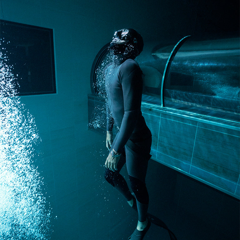 An underwater view of a diver in a sleek wetsuit near a pool structure, surrounded by rising bubbles. The diver wears a wristwatch, suggesting the watch’s underwater durability.