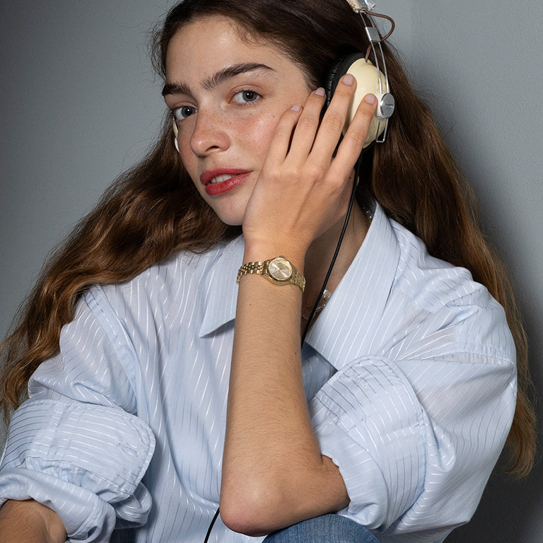 A young woman with long, wavy brown hair wearing vintage-style over-ear headphones and a light blue striped shirt. She rests her hand on her face, showcasing a gold wristwatch with a classic round dial on her wrist. She has a relaxed expression and looks directly at the camera.