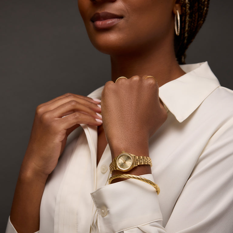 A close-up of a woman wearing a white blouse, accessorized with gold jewelry, including hoop earrings, rings, and a gold bracelet. She elegantly poses with her hand near her face, drawing attention to a gold wristwatch with a round dial on her wrist. The dark background contrasts with her outfit and accessories.