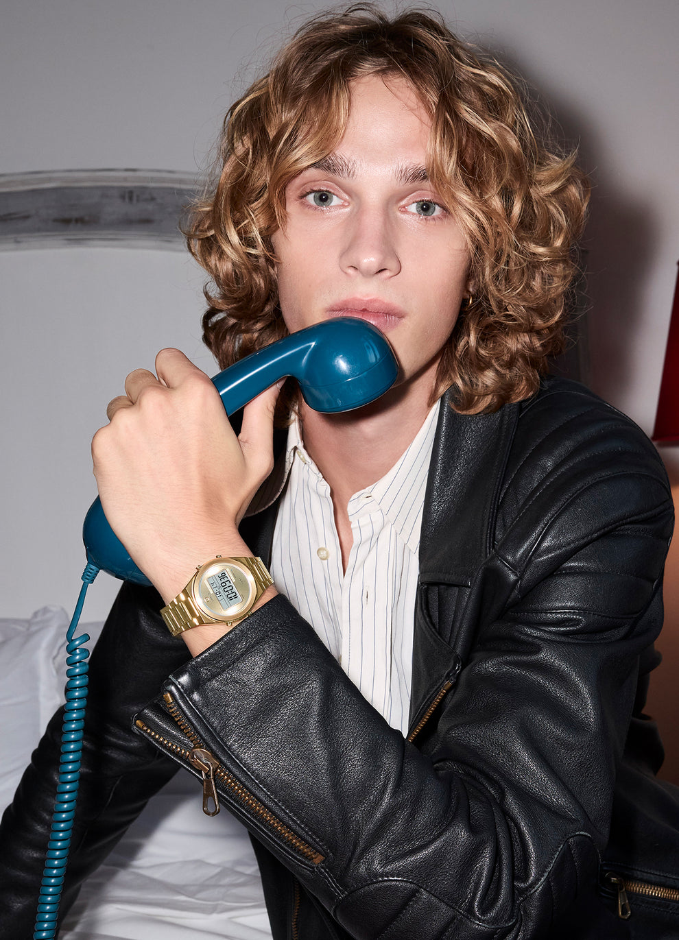A young man wearing a gold-tone Q Timex® Q80 Continental digital watch and a black leather jacket holds a blue rotary telephone receiver close to his face.