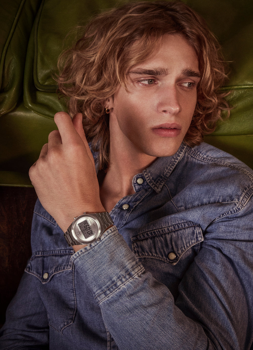 A young man with curly blonde hair and a leather jacket poses indoors, holding a retro blue telephone receiver to his mouth. He wears the gold-tone Q Timex® Q80 Continental digital watch on his wrist.