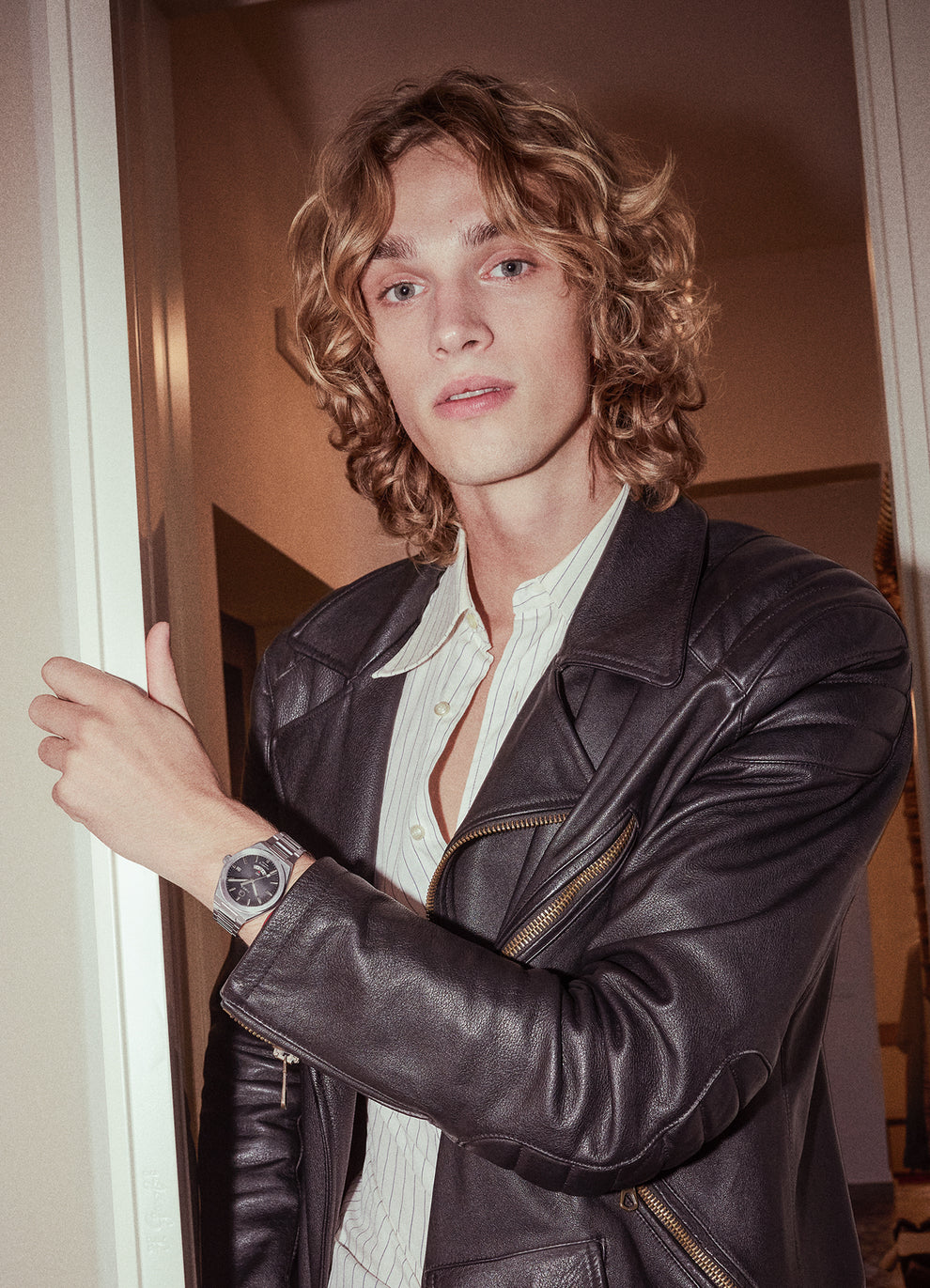 A person wearing the Q Timex® Continental GMT watch with a black dial and steel bracelet, styled with a leather jacket and open-collar shirt, standing in a doorway with soft indoor lighting.