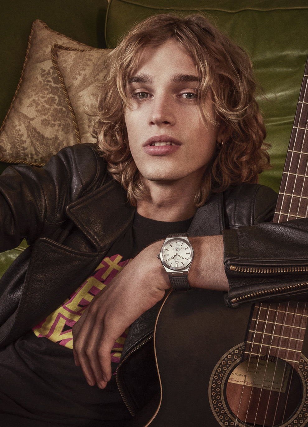 A young man with shoulder-length blonde hair lounges on a green leather couch wearing a leather jacket and the Q Timex® Continental GMT watch, paired with a graphic tee and acoustic guitar.