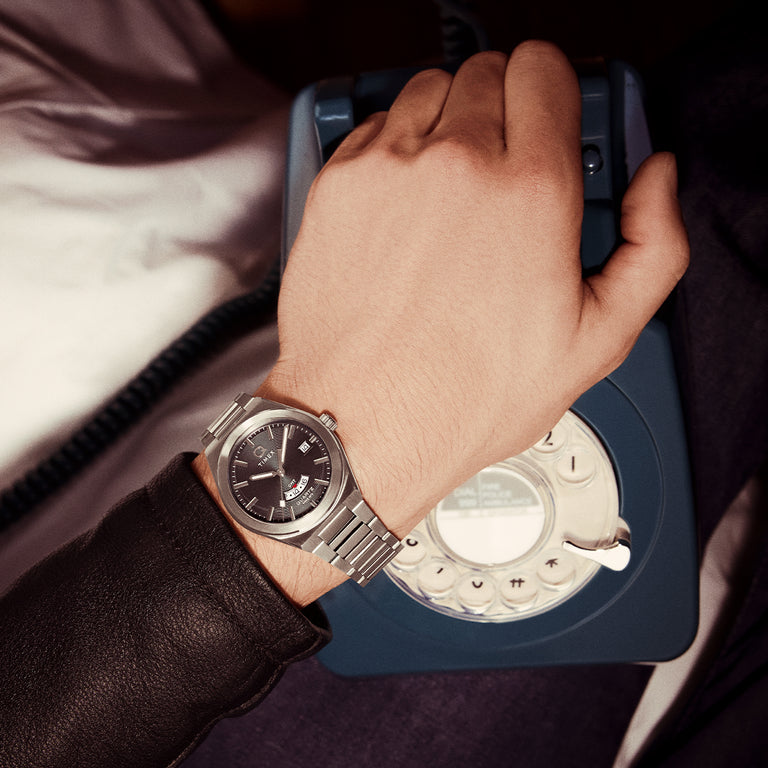 Close-up of the Q Timex® Continental GMT watch on a wrist, featuring its dual time zone function, black dial with red GMT scale, and brushed steel bracelet, while the wearer holds a vintage blue rotary phone.