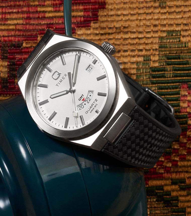Q Timex® Continental GMT watch with a silver dial and black woven-texture strap, draped over a vintage teal rotary phone, with a warm tapestry textile in the background.